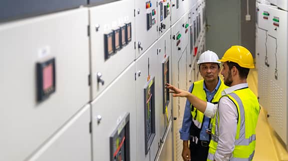 Two men wearing hard hats examine electrical panels, engaged in a technical discussion in an industrial environment.