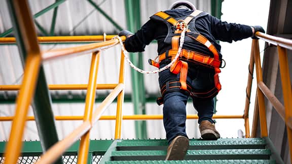 A man in a safety harness climbs a set of stairs, demonstrating safety measures in a construction or industrial setting.