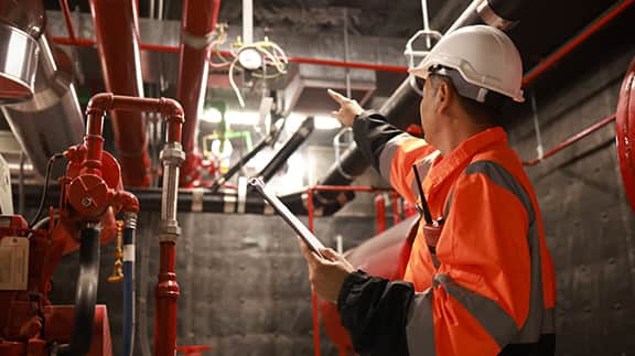 A man wearing an orange safety vest holds a clipboard, standing in a work environment.