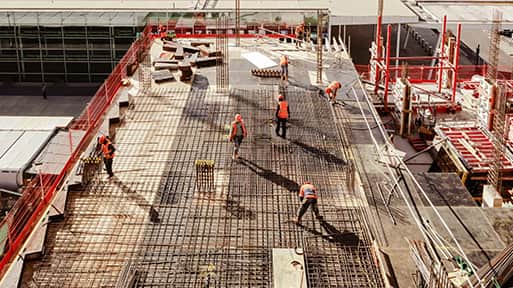 Construction workers in orange vests work on a building site, laying down metal rebar on a concrete structure.